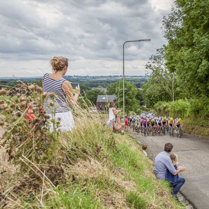 Peloton met wielrenners fietst een berg op met publiek in de hoge berm.
